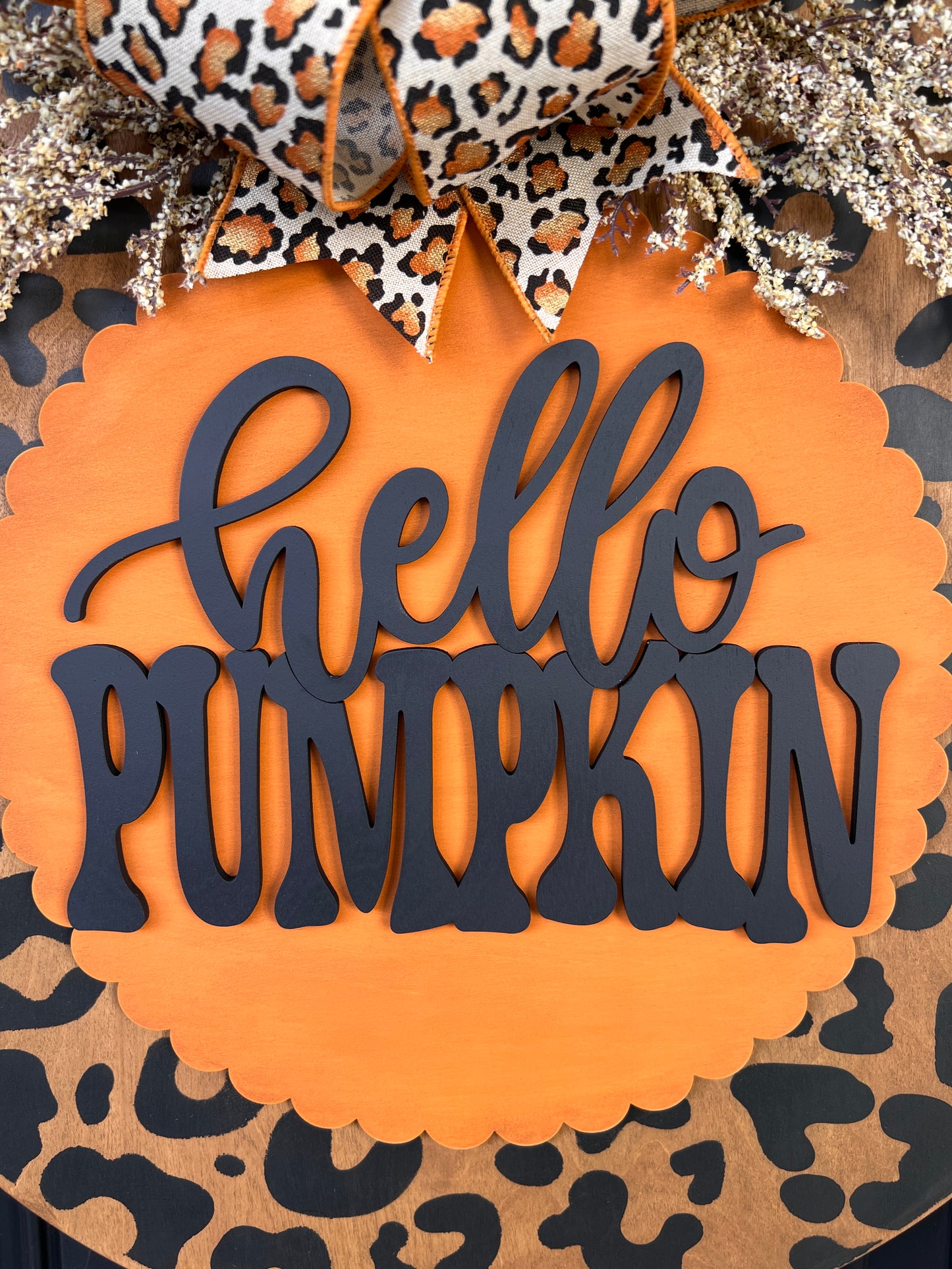 Hello Pumpkin leopard print fall door hanger with layered wood lettering, designer bow, seasonal greenery, and woodgrain animal print background displayed on a front door