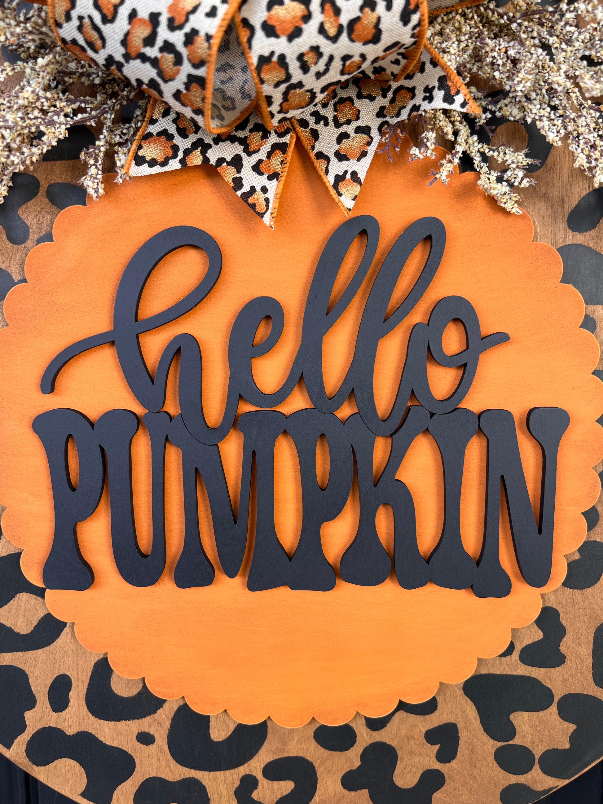 Hello Pumpkin leopard print fall door hanger with layered wood lettering, designer bow, seasonal greenery, and woodgrain animal print background displayed on a front door
