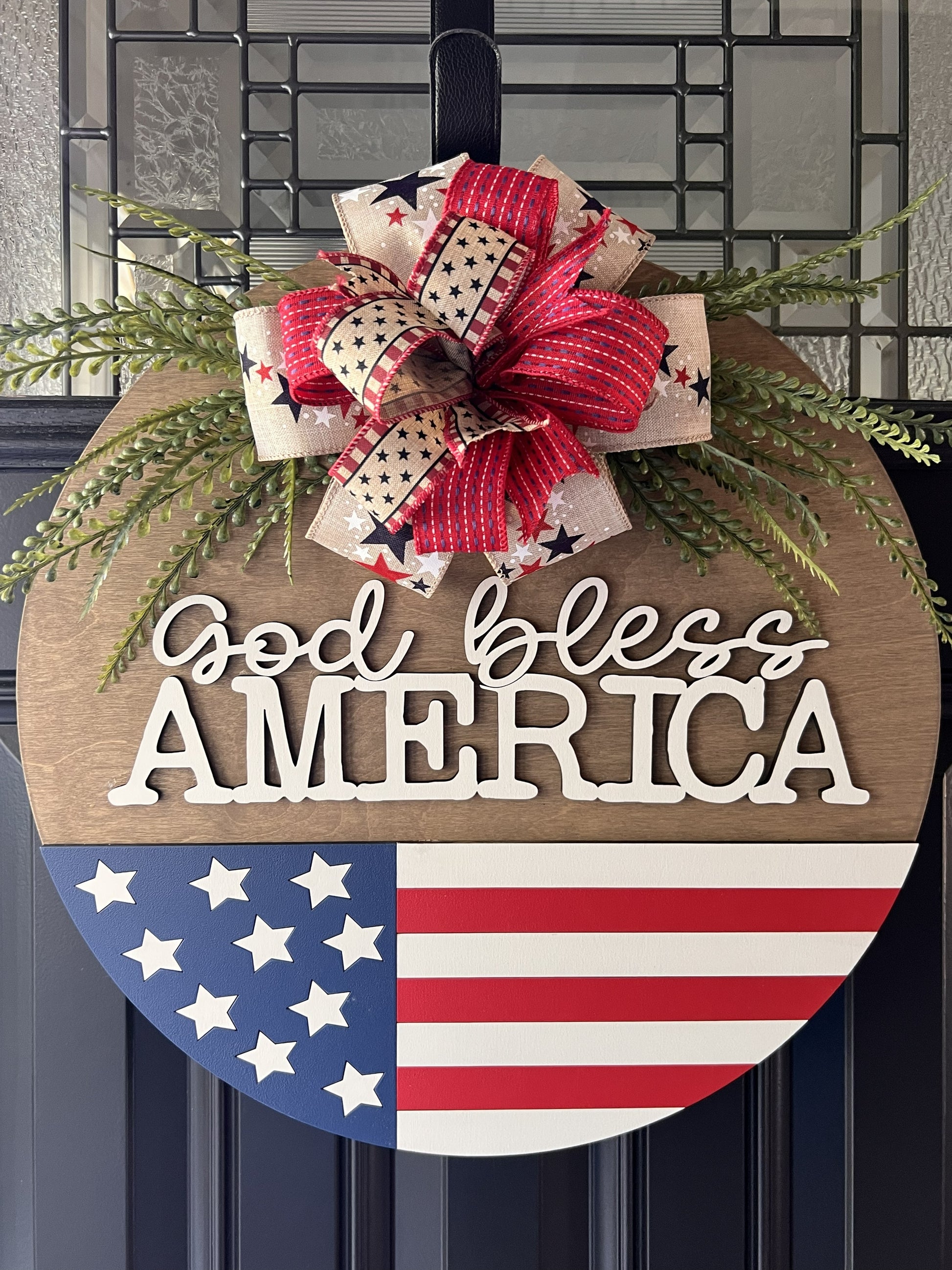 Decorative sign with 'God bless America' text and American flag design, featuring a bow and beaded fern greenery