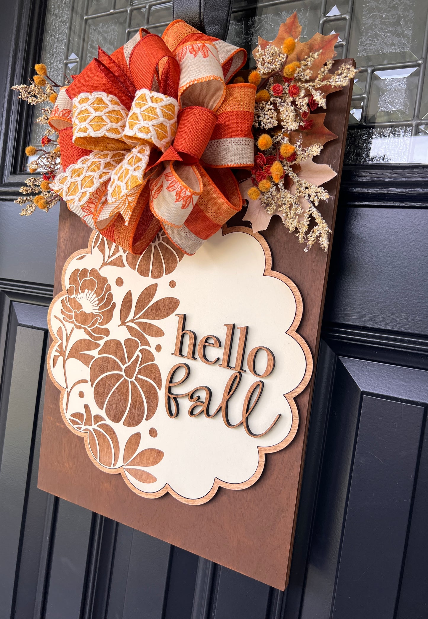 An autumn-themed wooden door wreath hanging on a black paneled front door. The wreath features a large orange and beige patterned ribbon bow at the top, dried leaves, small orange berries, and a scalloped cream sign with the words 'hello fall' in brown script.