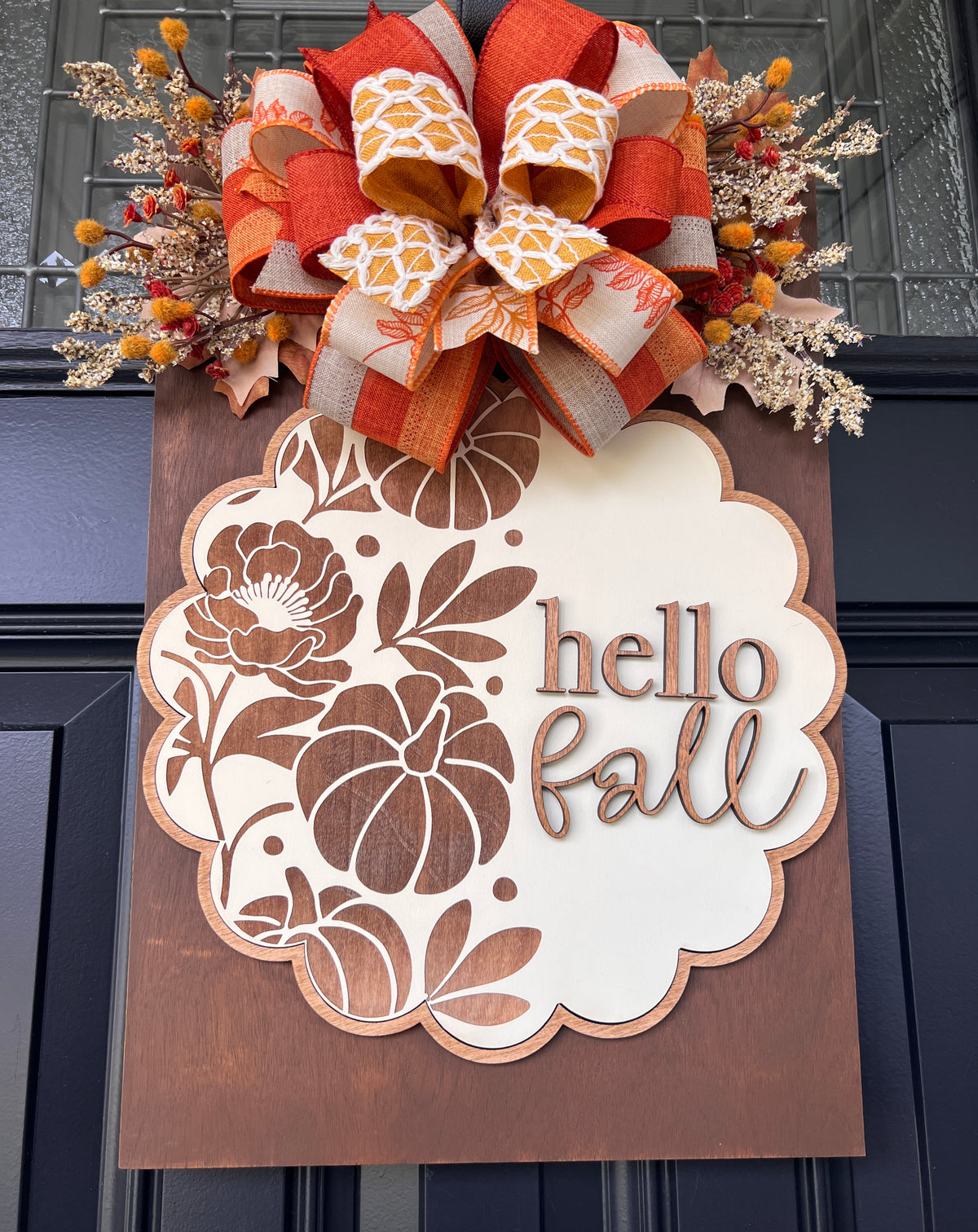 An autumn-themed wooden door sign with a scalloped white panel reading 'hello fall' in brown script and floral cutout designs, mounted on a dark door. A large decorative bow in orange, beige, and white patterned ribbon sits above the sign, with fall foliage accents on both sides.