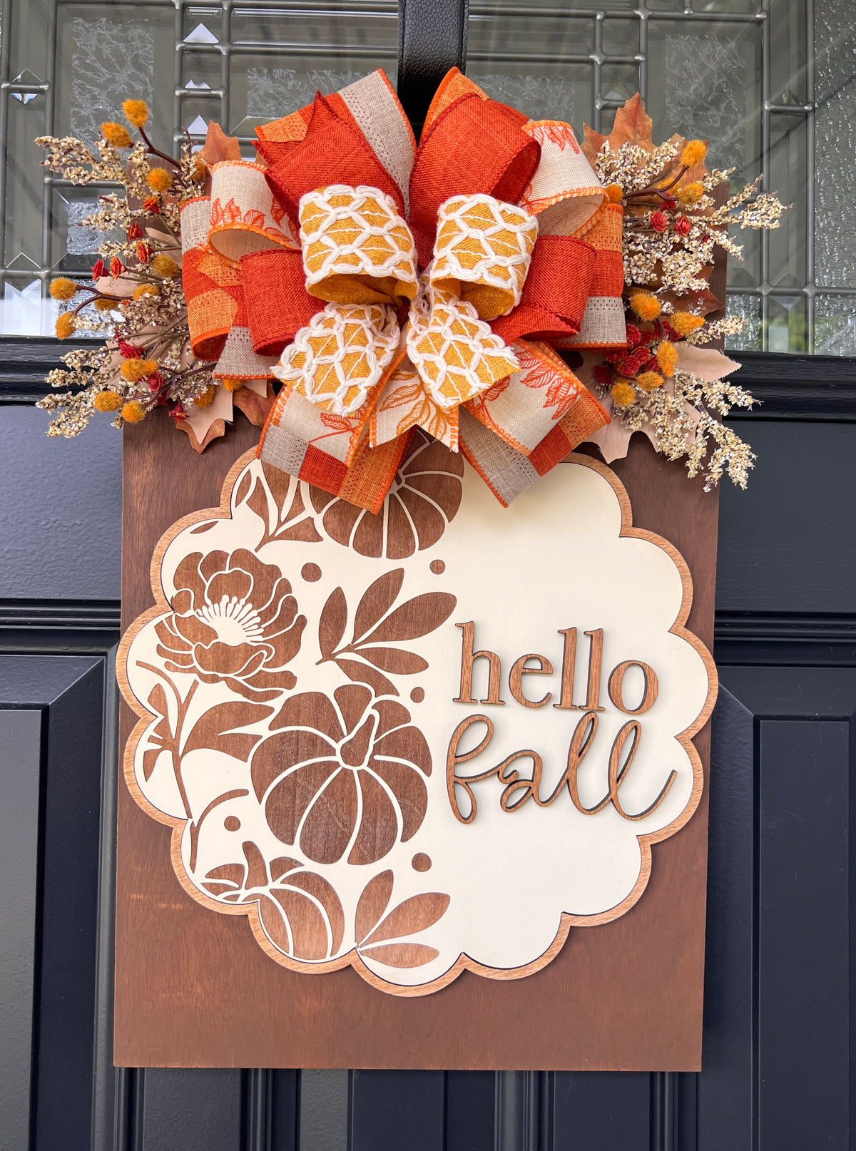 Hello Fall engraved scalloped rectangle door hanger - 12x18 warm stained birchwood base with cream laser-engraved scalloped oval center featuring pumpkin and floral design, topped with layered burnt orange cream floral and mustard geometric ribbon bow with dried fall berry sprays on black front door