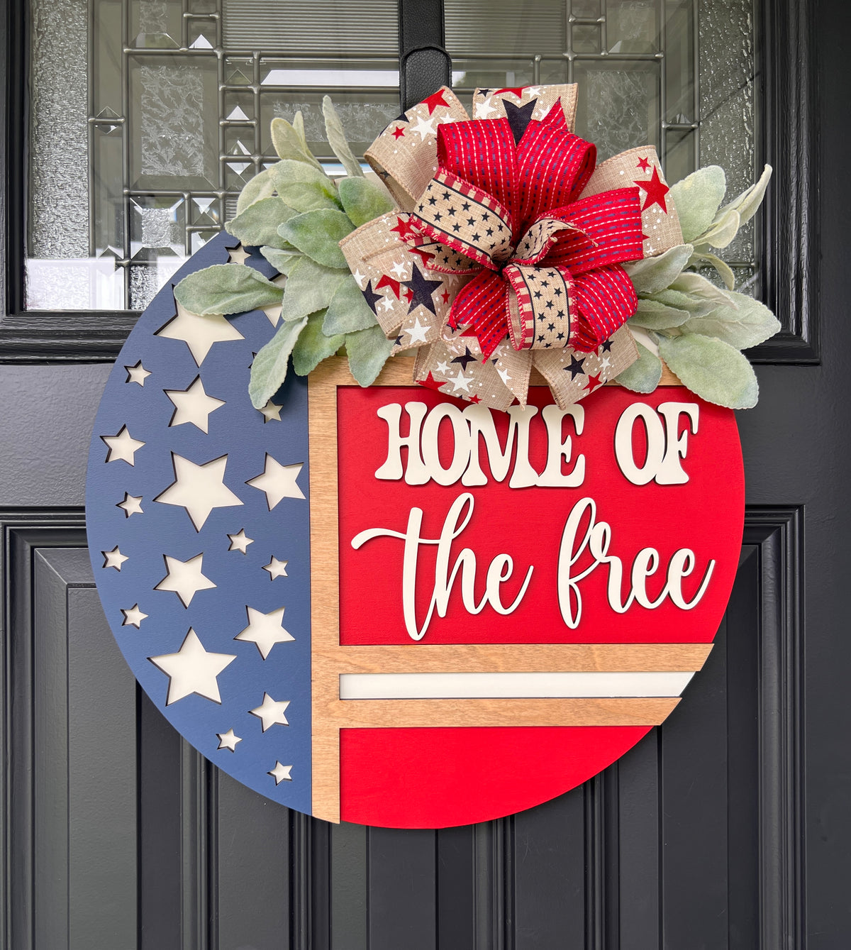 Home of the Free patriotic door hanger - red version with laser-cut star field on navy left panel, bold 3D cream lettering on red right panel, layered red stripe and star print ribbon bow with lambs ear greenery on black front door