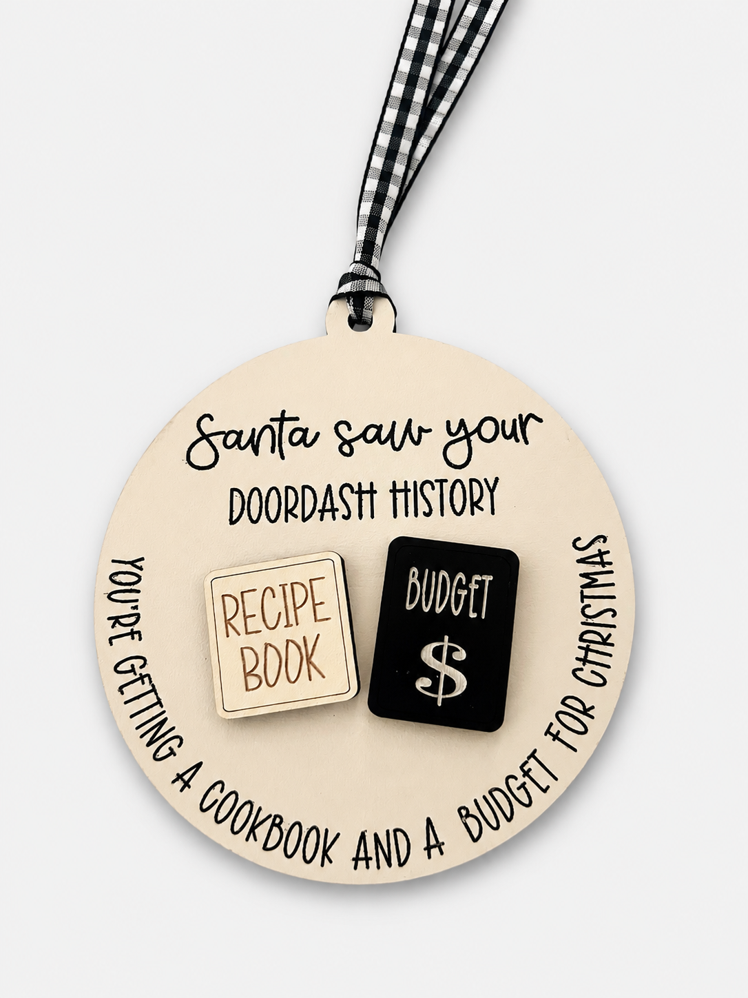 A round beige ornament with a black-and-white gingham ribbon, featuring printed text that reads 'Santa saw your doordash history' and two small icon-like blocks reading 'RECIPE BOOK' in beige and 'BUDGET $' in black.