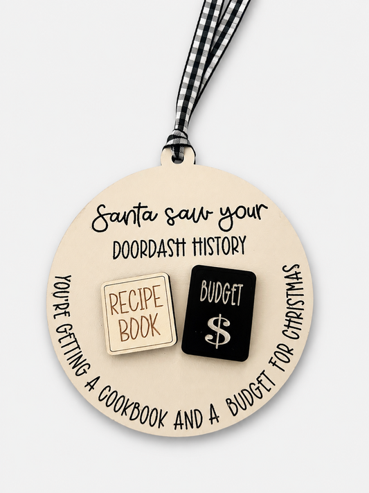 A round beige ornament with a black-and-white gingham ribbon, featuring printed text that reads 'Santa saw your doordash history' and two small icon-like blocks reading 'RECIPE BOOK' in beige and 'BUDGET $' in black.
