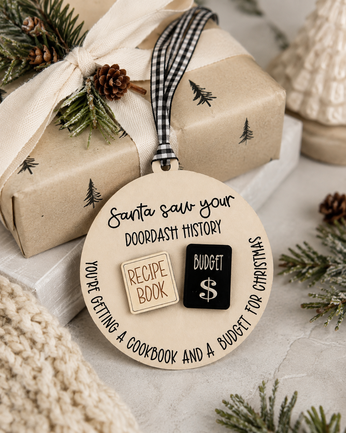 A round wooden Christmas ornament with a black-and-white gingham ribbon, displayed on top of a wrapped gift box. The ornament features a natural wood background with black text reading 'Santa saw your doordash history' and two small decorative pieces: a beige wood-grain 'Recipe Book' tab and a black wood-grain 'Budget $' tab.