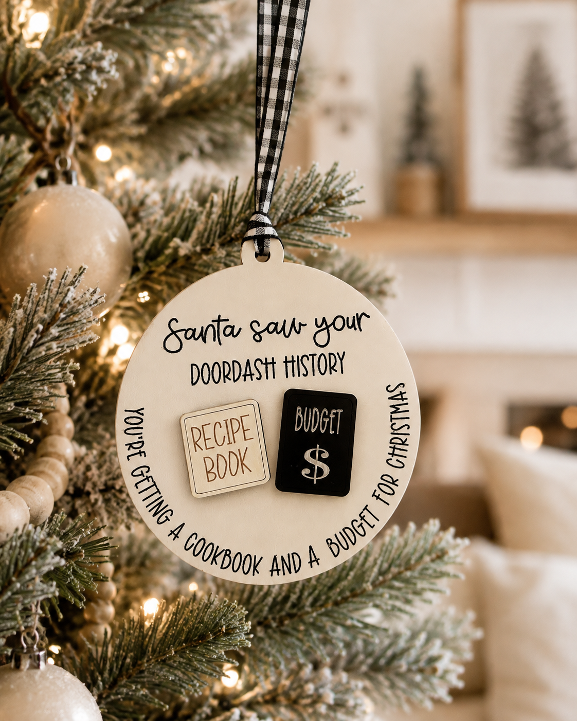 A round Christmas ornament hanging from a black-and-white gingham ribbon on a decorated tree. The ornament has printed text reading 'Santa saw your DOODASH HISTORY' and 'YOU'RE GETTING A COOKBOOK AND A BUDGET FOR CHRISTMAS,' plus two small coin-like inserts labeled 'RECIPE BOOK' and 'BUDGET $'.