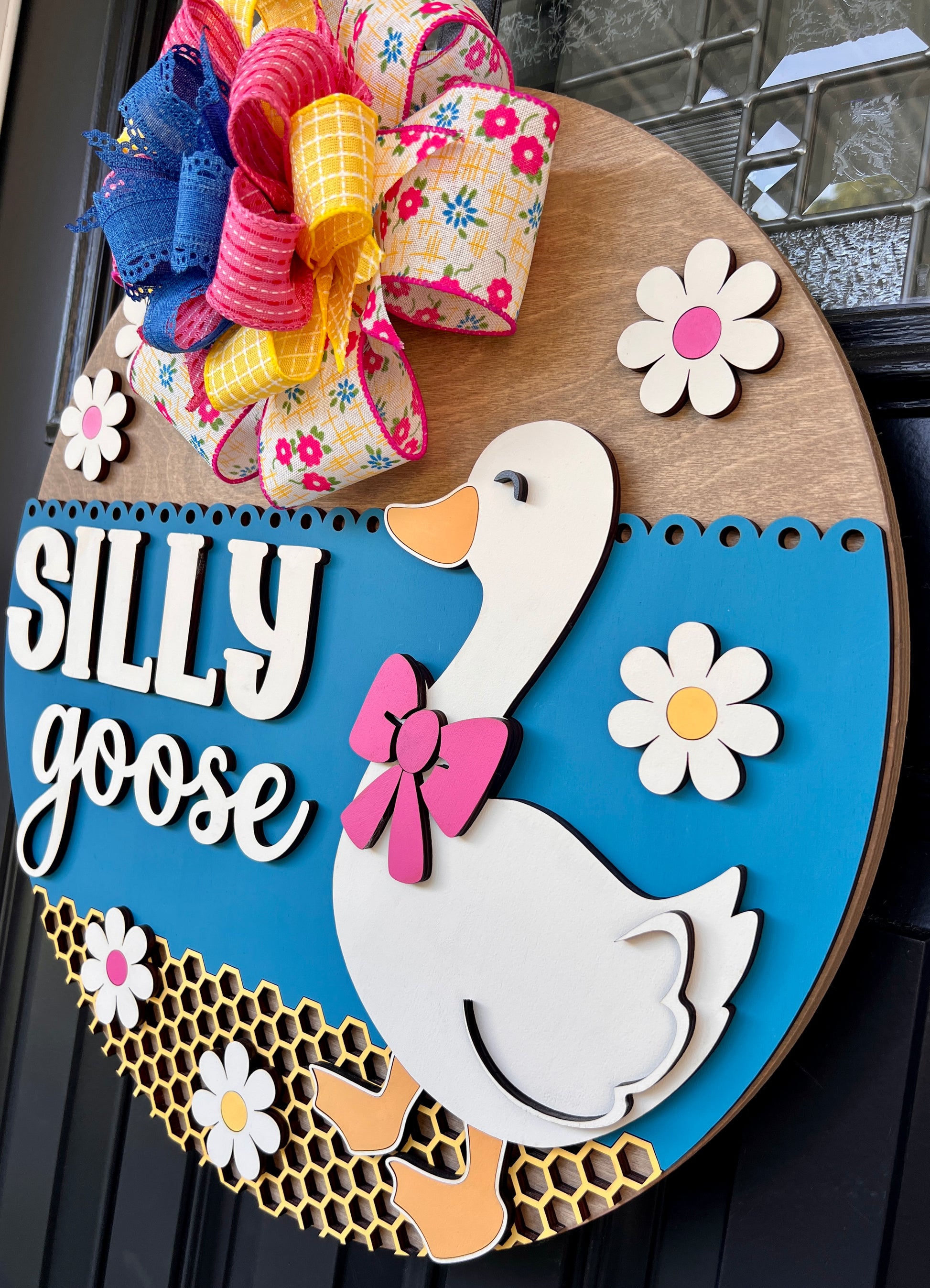 Handcrafted Silly Goose door hanger featuring a white goose with a pink bow, layered 3D lettering, colorful daisy accents, blue background, honeycomb detail, and decorative bow for farmhouse front door decor.