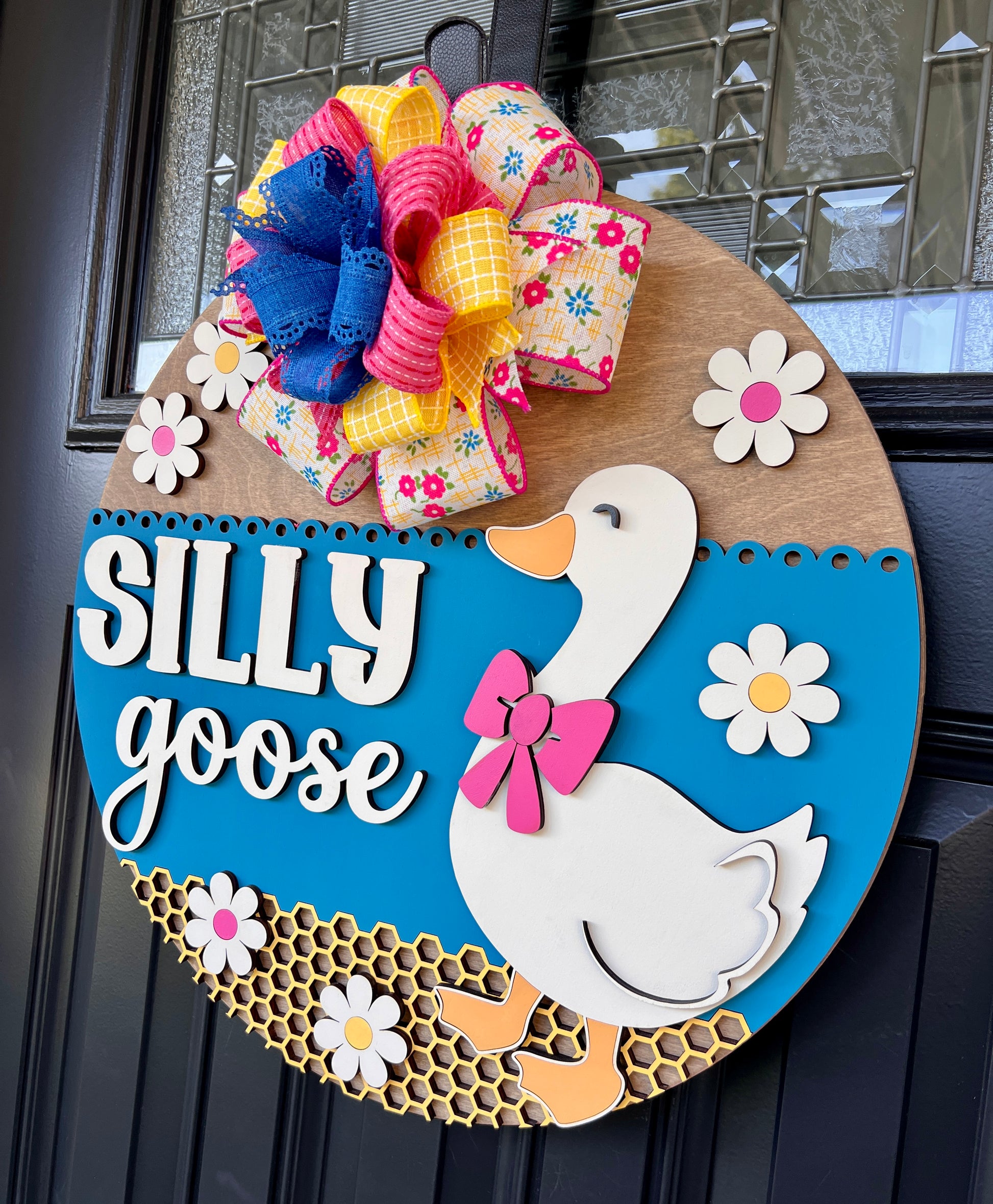 Handcrafted Silly Goose door hanger featuring a white goose with a pink bow, layered 3D lettering, colorful daisy accents, blue background, honeycomb detail, and decorative bow for farmhouse front door decor.