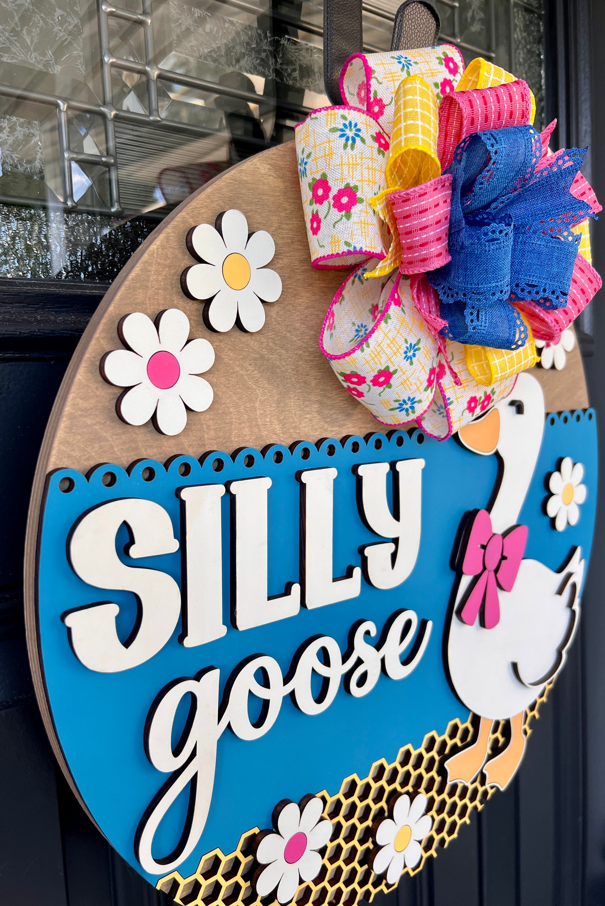 Handcrafted Silly Goose door hanger featuring a white goose with a pink bow, layered 3D lettering, colorful daisy accents, blue background, honeycomb detail, and decorative bow for farmhouse front door decor.