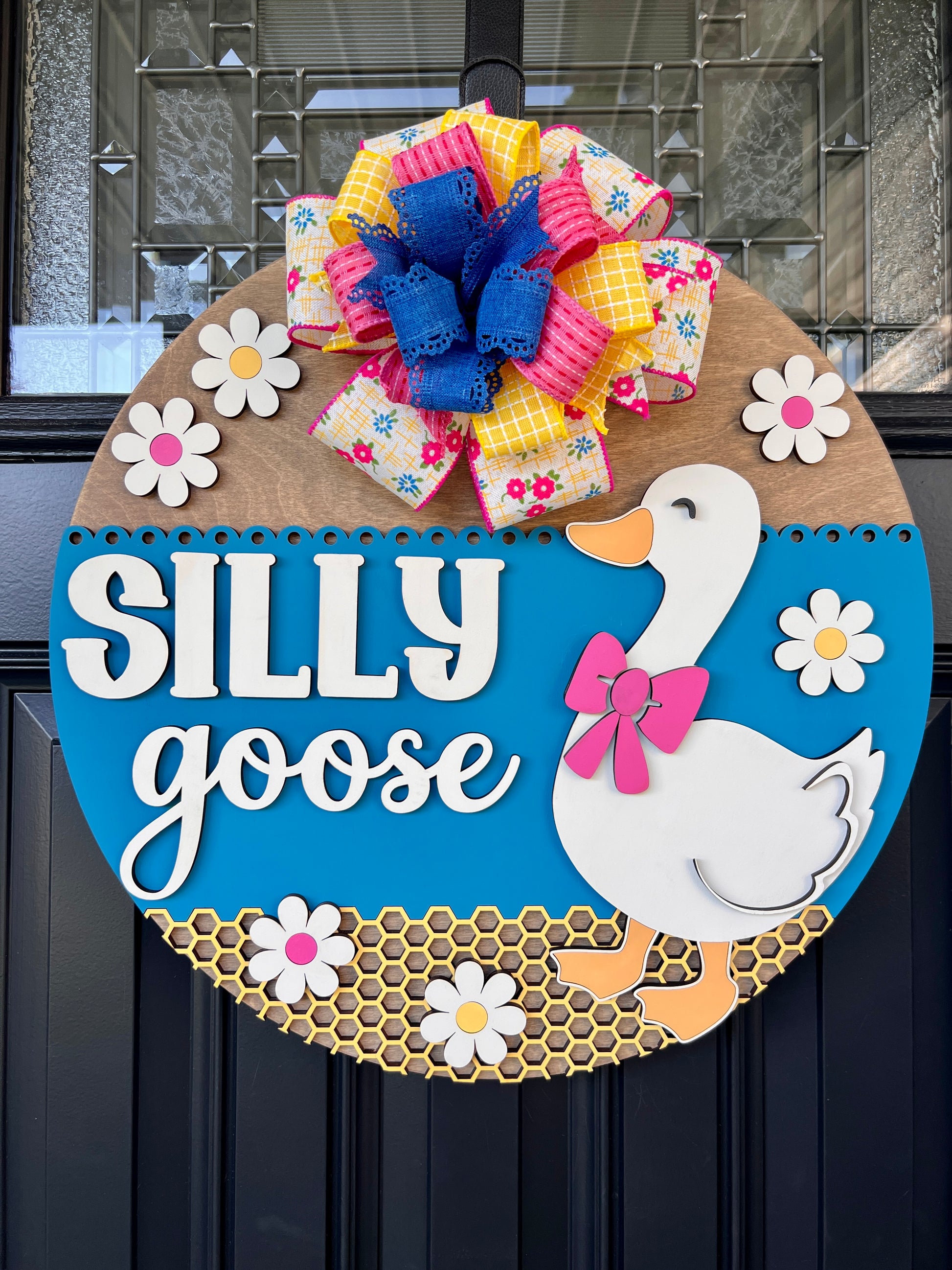 Handcrafted Silly Goose door hanger featuring a white goose with a pink bow, layered 3D lettering, colorful daisy accents, blue background, honeycomb detail, and decorative bow for farmhouse front door decor.