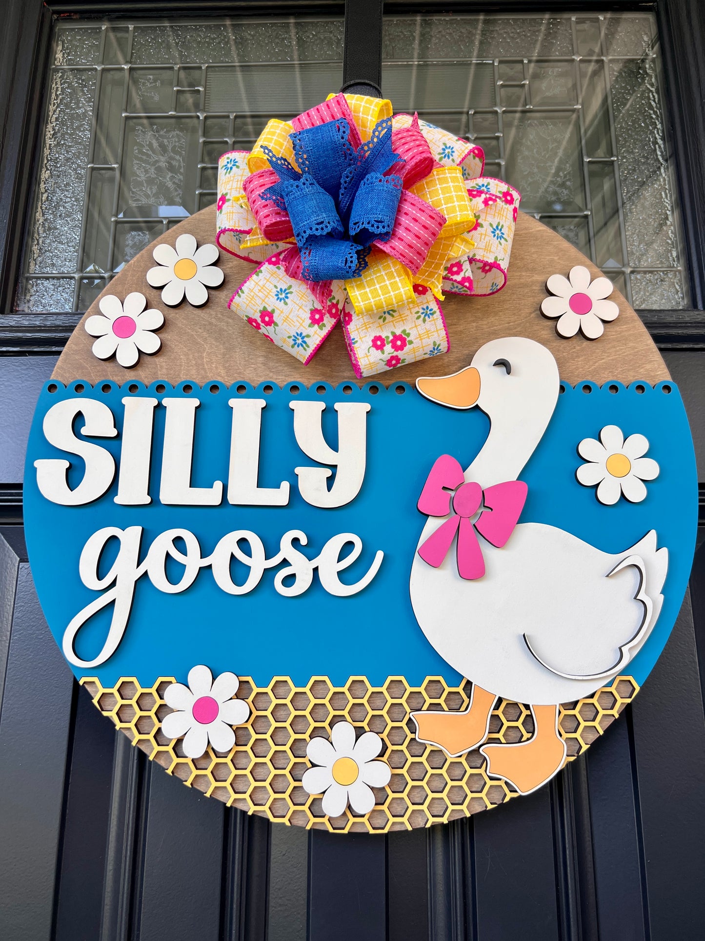 Handcrafted Silly Goose door hanger featuring a white goose with a pink bow, layered 3D lettering, colorful daisy accents, blue background, honeycomb detail, and decorative bow for farmhouse front door decor.