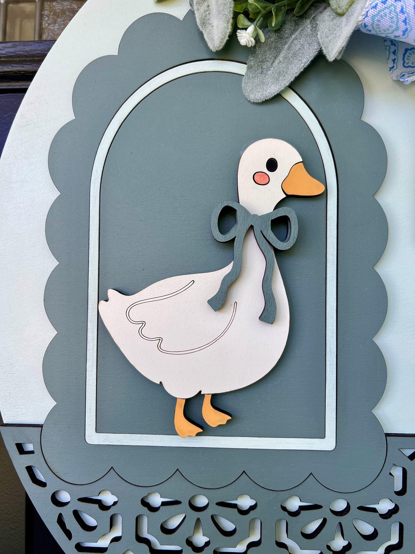 A decorative wooden wall hanging featuring a stylized white goose with a gray bow, orange beak and feet, set against a gray arched panel with a scalloped border.