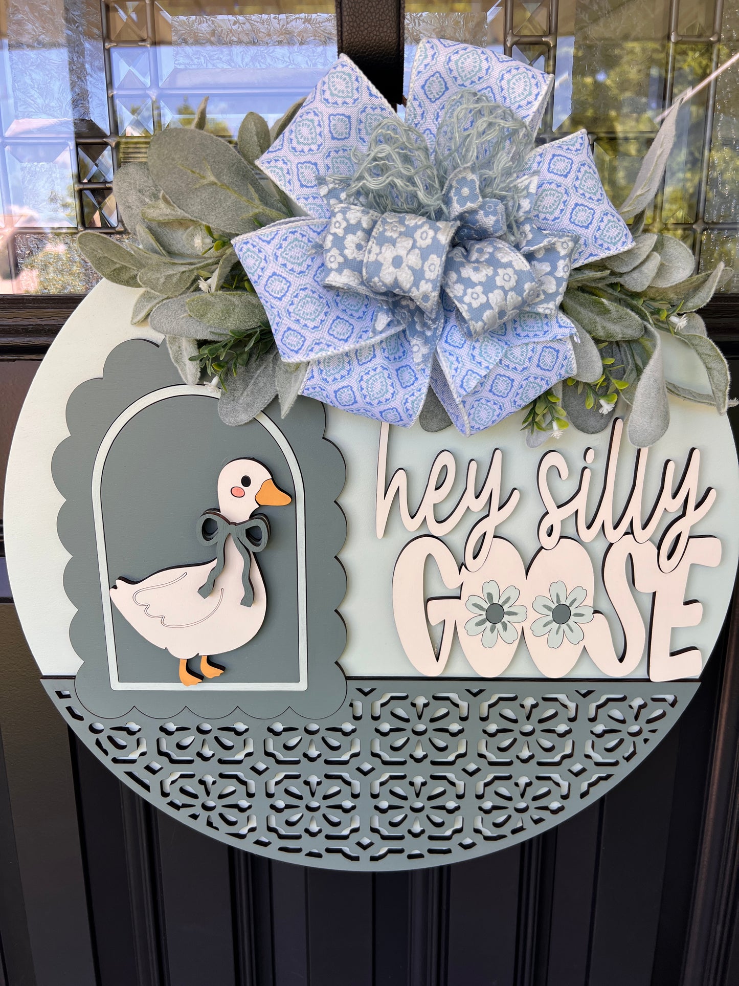A decorative circular wreath hanging on a dark door, featuring a large blue patterned bow, green foliage, a white goose cutout with an orange beak, and the phrase “hey silly GOOSE” in layered white and pale blue lettering.