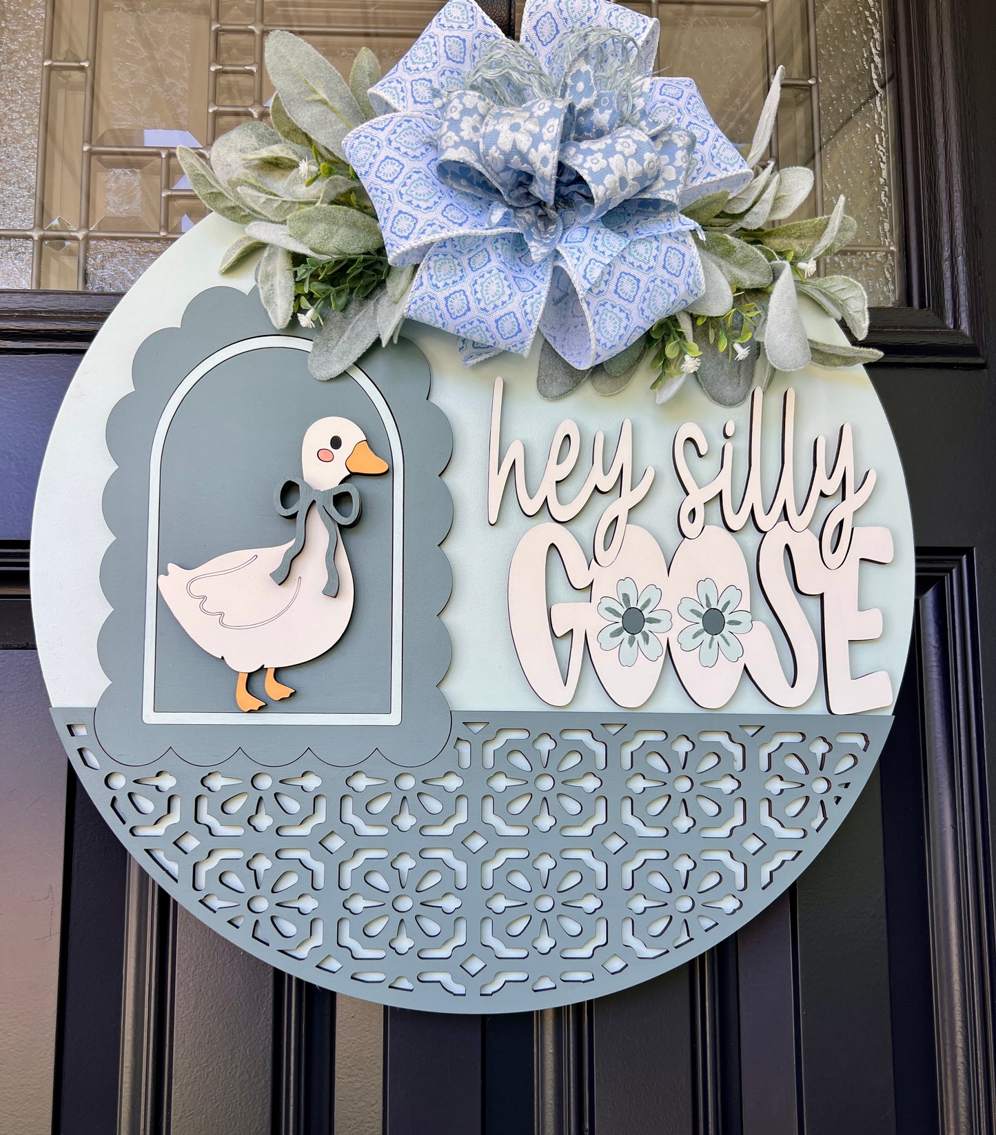 A decorative circular door wreath hanging on a dark front door. The wreath is light blue with a large blue-and-white patterned bow and greenery at the top, a white goose cutout on the left, and the phrase “hey silly GOOSE” in white lettering on the right.