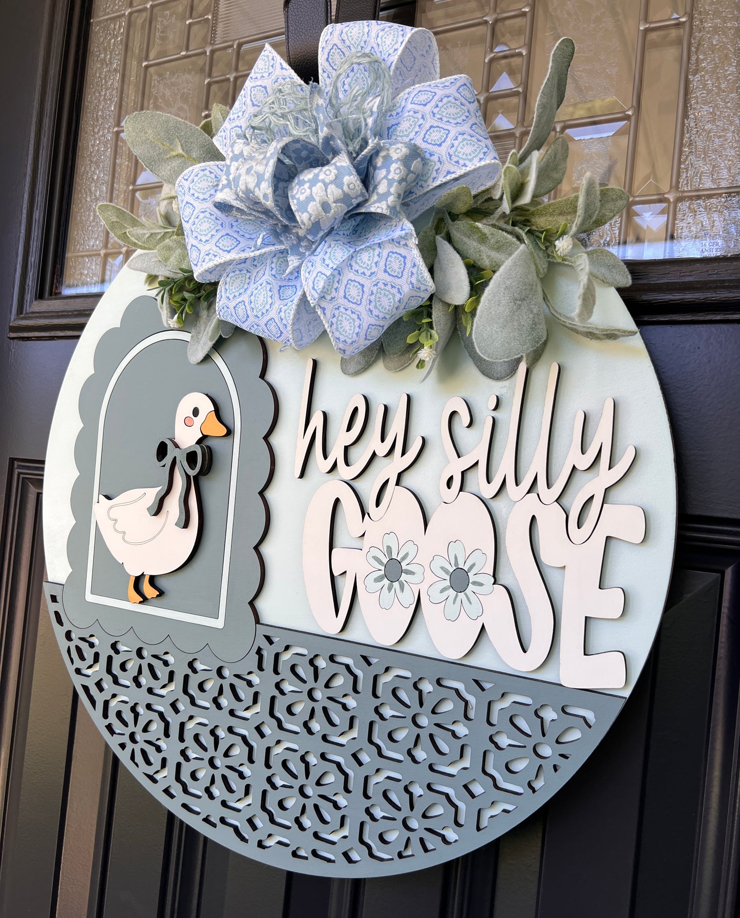 A decorative oval wreath hanging on a dark front door. The wreath has a large blue patterned bow at the top, green faux foliage, a white goose cutout with a black bow, and the phrase “hey silly GOSE” in white lettering over a blue patterned lower half with floral cutout detailing.