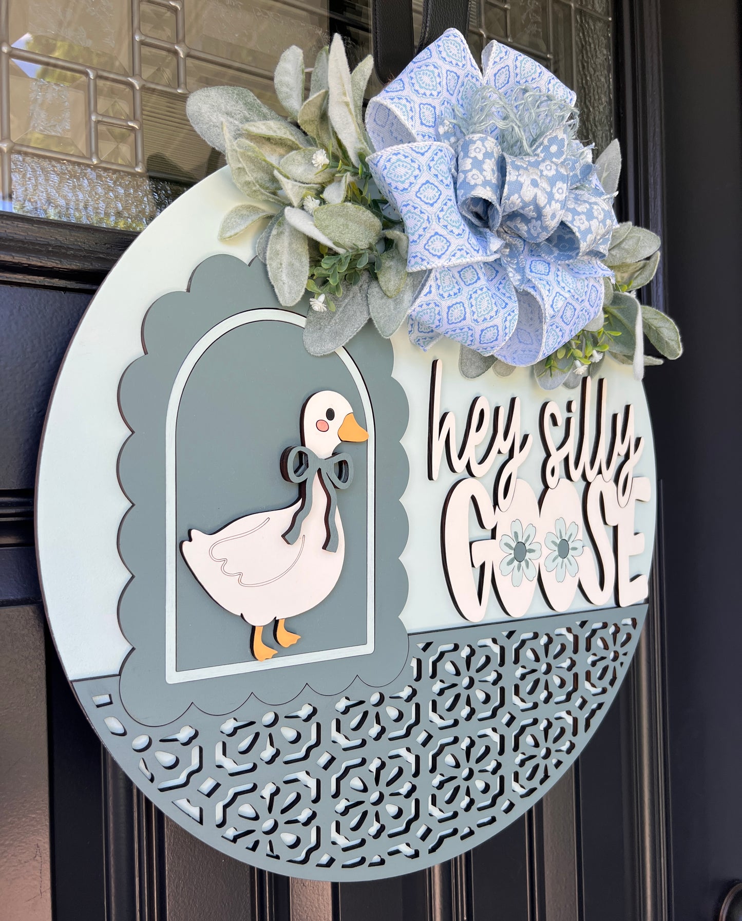 A decorative oval wreath hanging on a dark front door. The wreath has a light blue and white patterned bow, faux greenery, a white duck cutout, and the phrase “hey silly GOSE” in white script over a gray background.