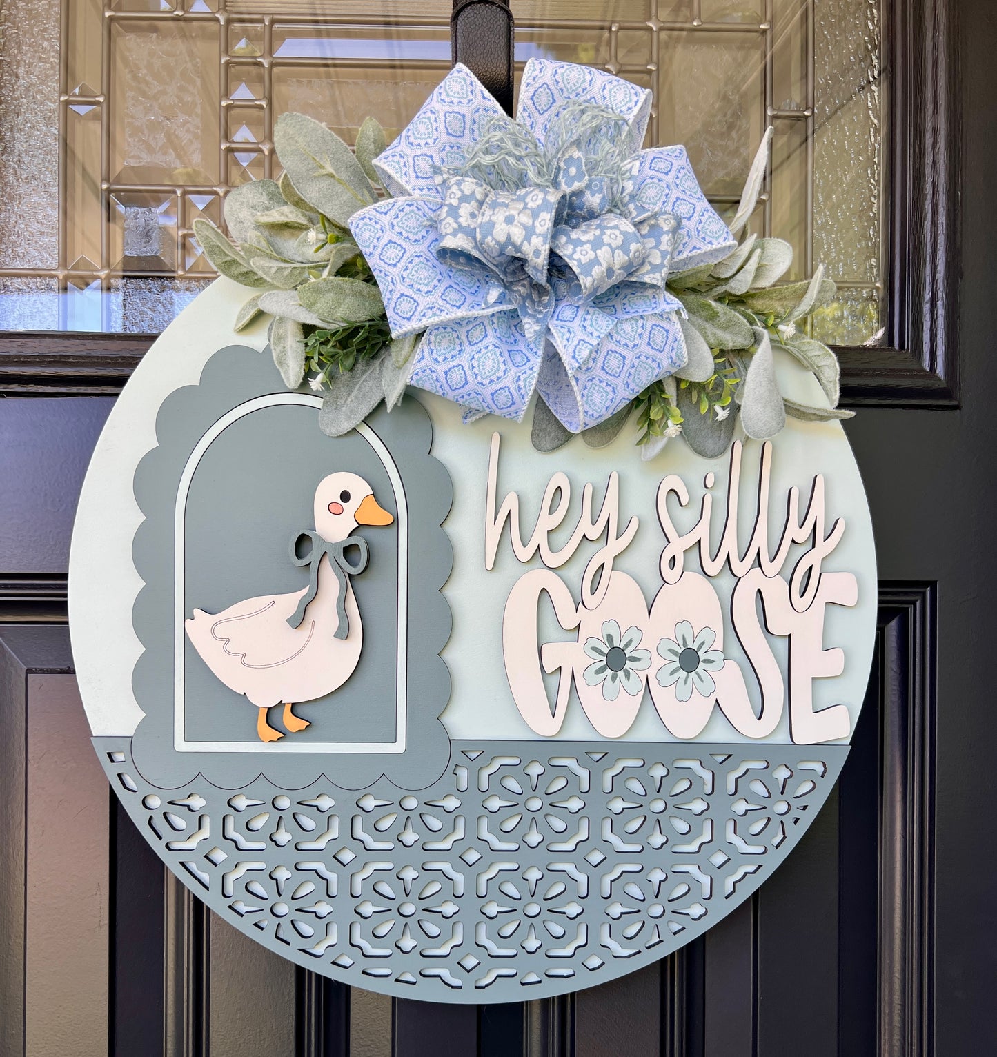 A decorative round wooden door wreath hanging on a dark door. The wreath features a light blue and white patterned bow at the top, a white goose illustration in a scalloped frame, and the phrase 'hey silly GOOSE' in layered white and pale blue lettering.