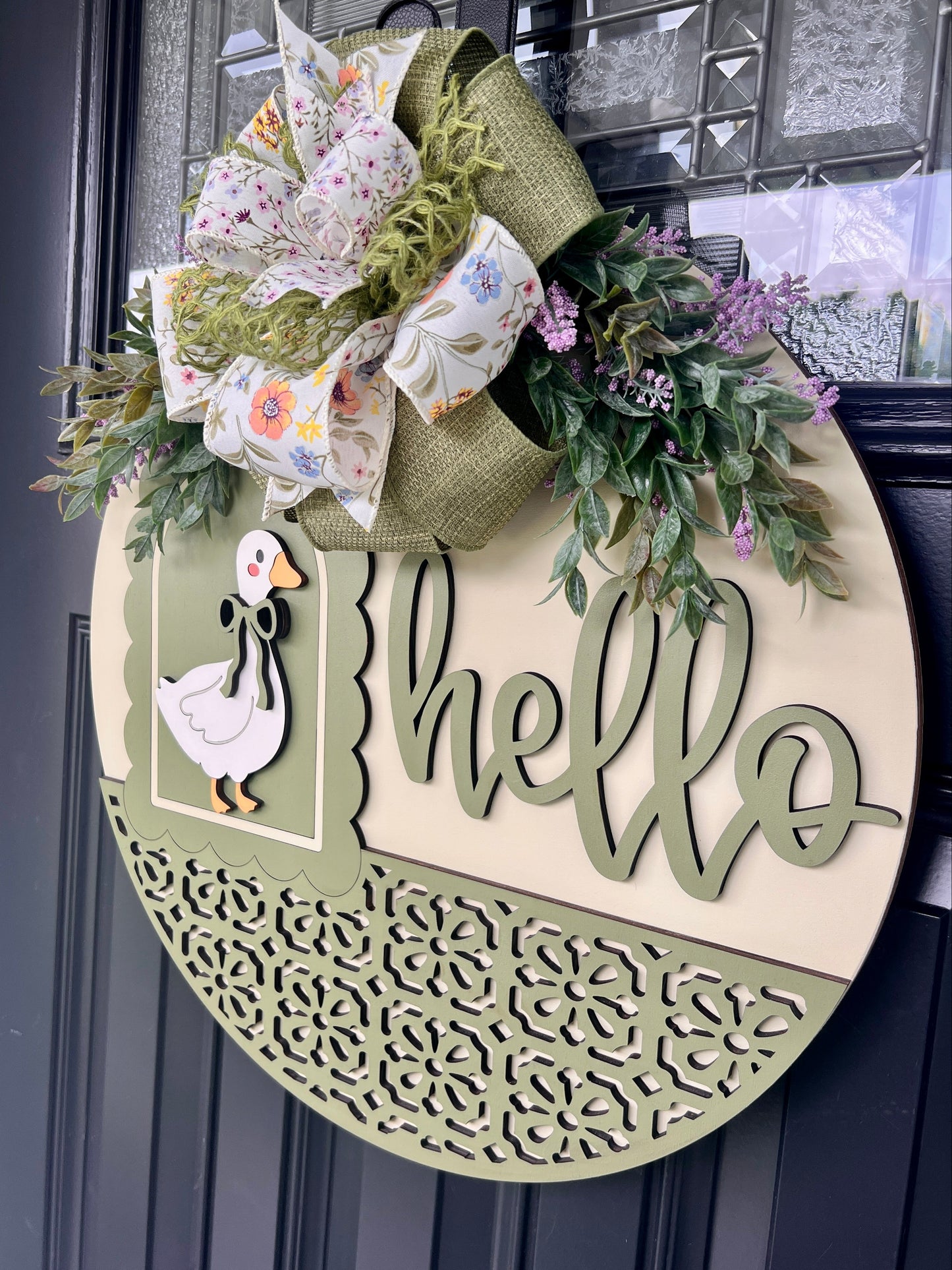 Angled view of a handmade wooden Hello goose door hanger showcasing dimensional laser-cut lettering, sage green frame, decorative scrollwork, and spring floral bow.