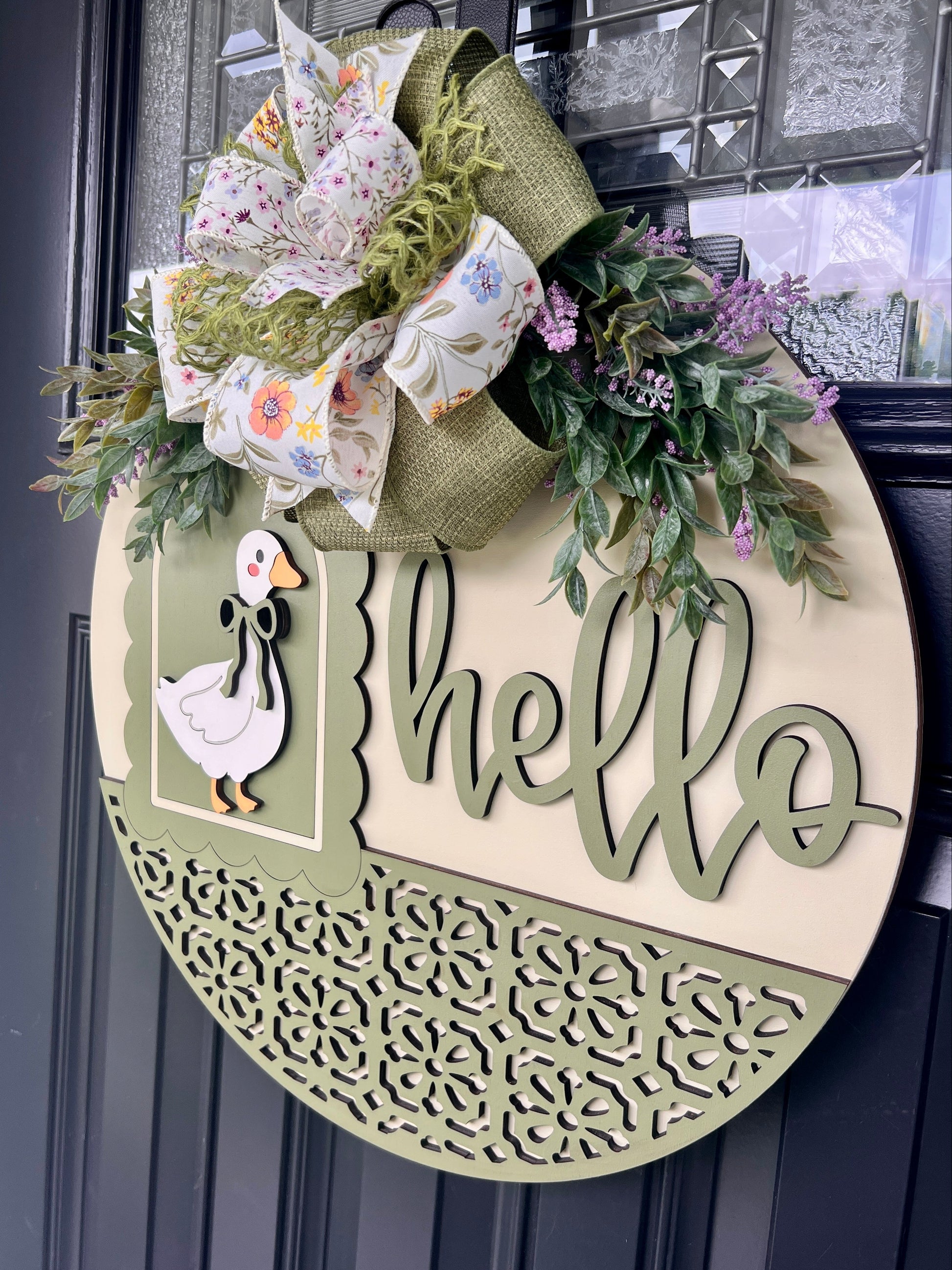 Angled view of a handmade wooden Hello goose door hanger showcasing dimensional laser-cut lettering, sage green frame, decorative scrollwork, and spring floral bow.