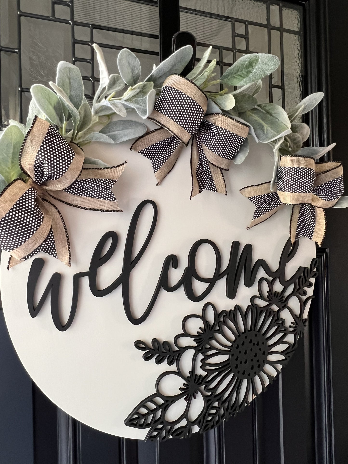 Decorative wreath with 'welcome' text and floral design- lambsear greeery and bows on a black door.