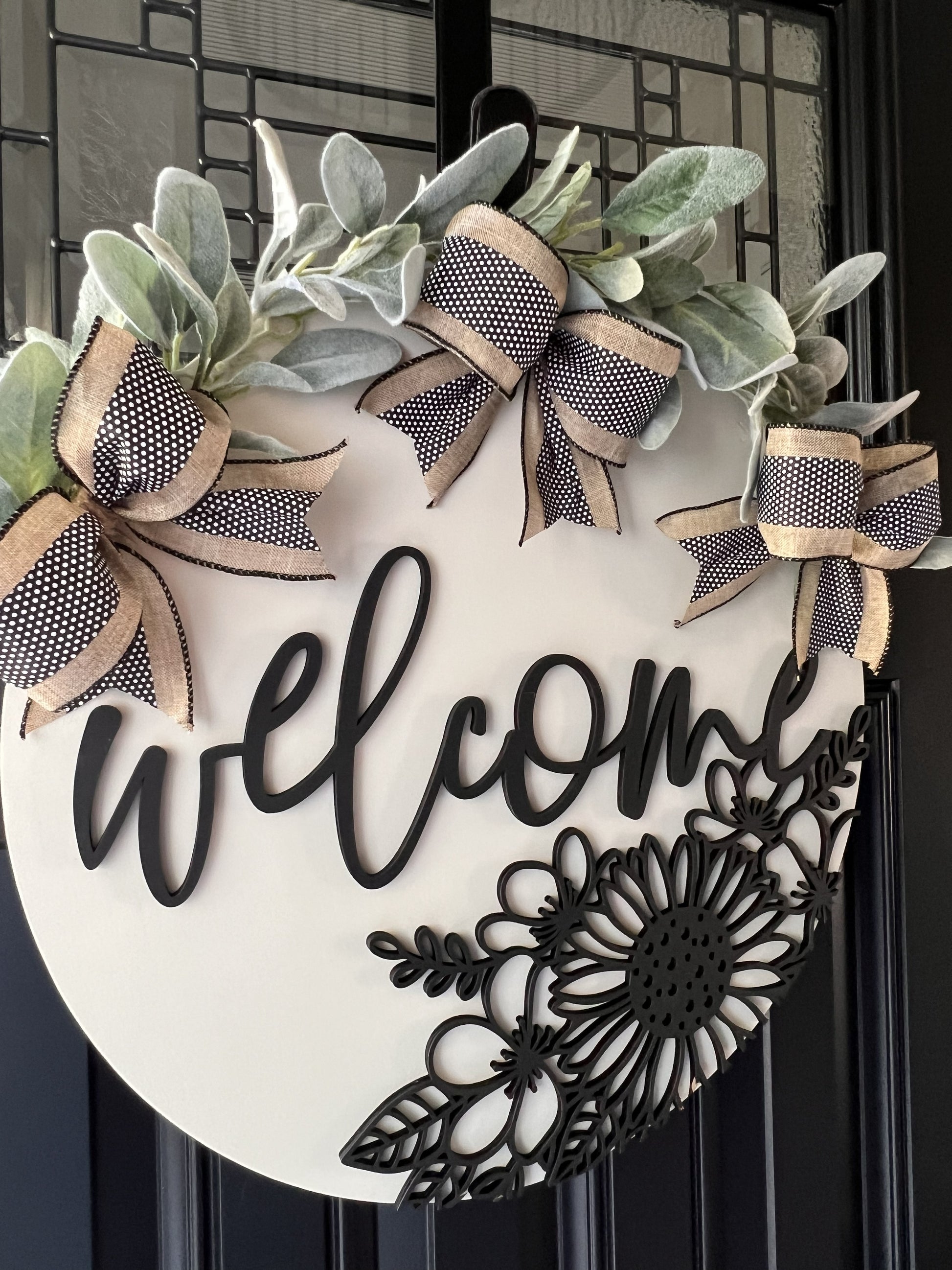 Decorative wreath with 'welcome' text and floral design- lambsear greeery and bows on a black door.