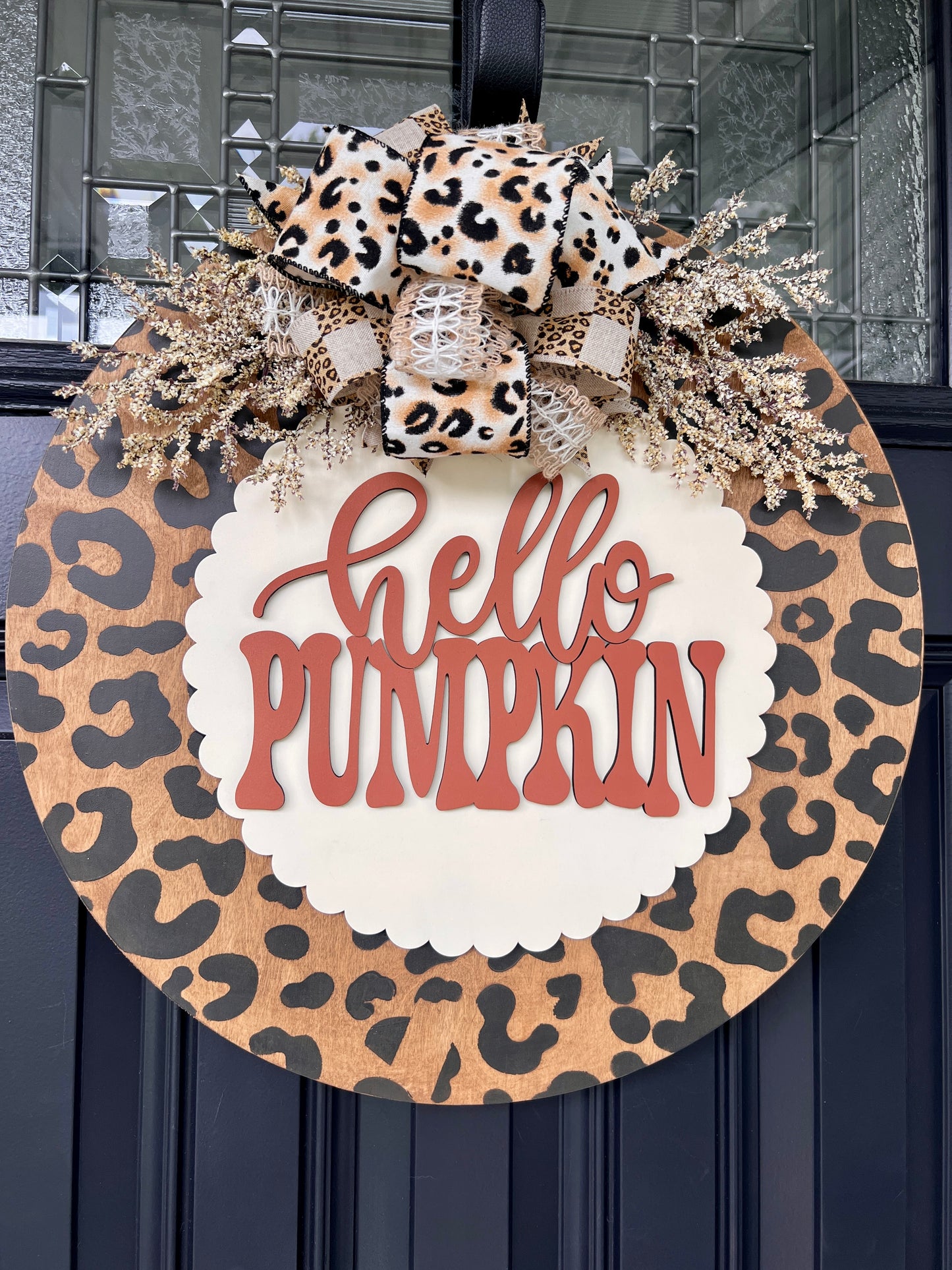 Hello Pumpkin leopard print fall door hanger with layered wood lettering, designer bow, seasonal greenery, and woodgrain animal print background displayed on a front door