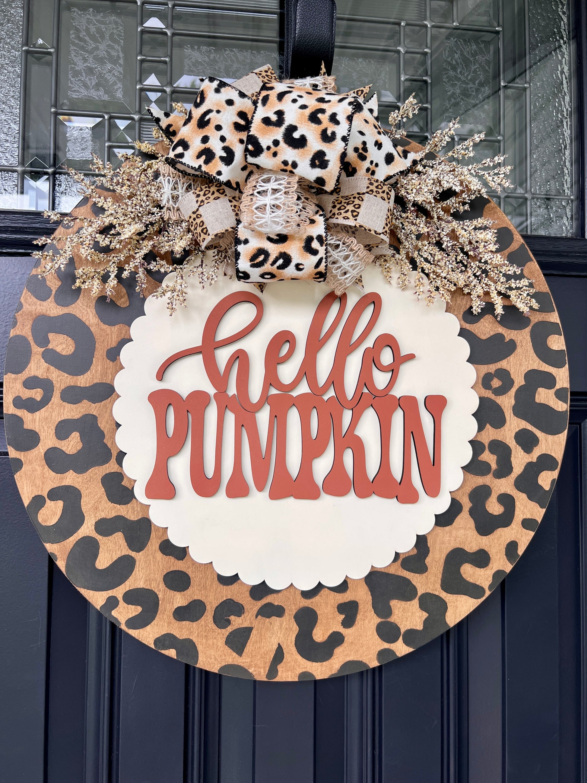 Hello Pumpkin leopard print fall door hanger with layered wood lettering, designer bow, seasonal greenery, and woodgrain animal print background displayed on a front door