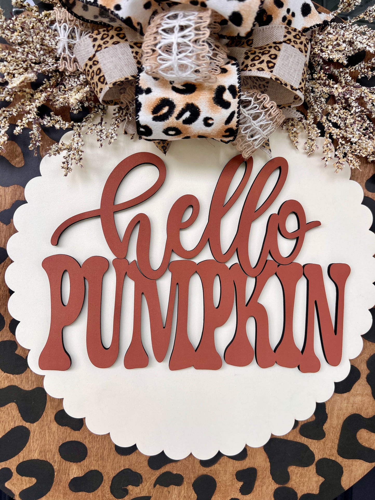 Hello Pumpkin leopard print fall door hanger with layered wood lettering, designer bow, seasonal greenery, and woodgrain animal print background displayed on a front door