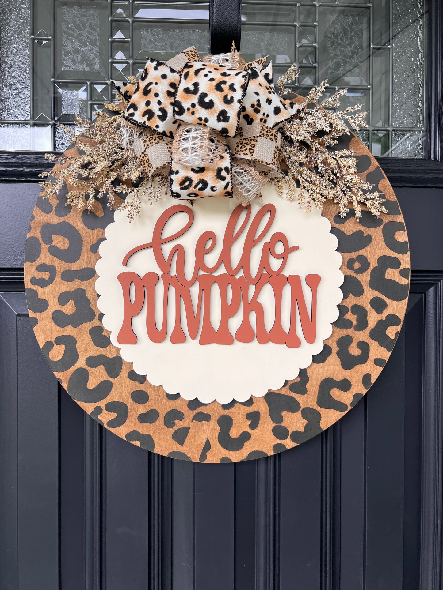 Hello Pumpkin leopard print fall door hanger with layered wood lettering, designer bow, seasonal greenery, and woodgrain animal print background displayed on a front door
