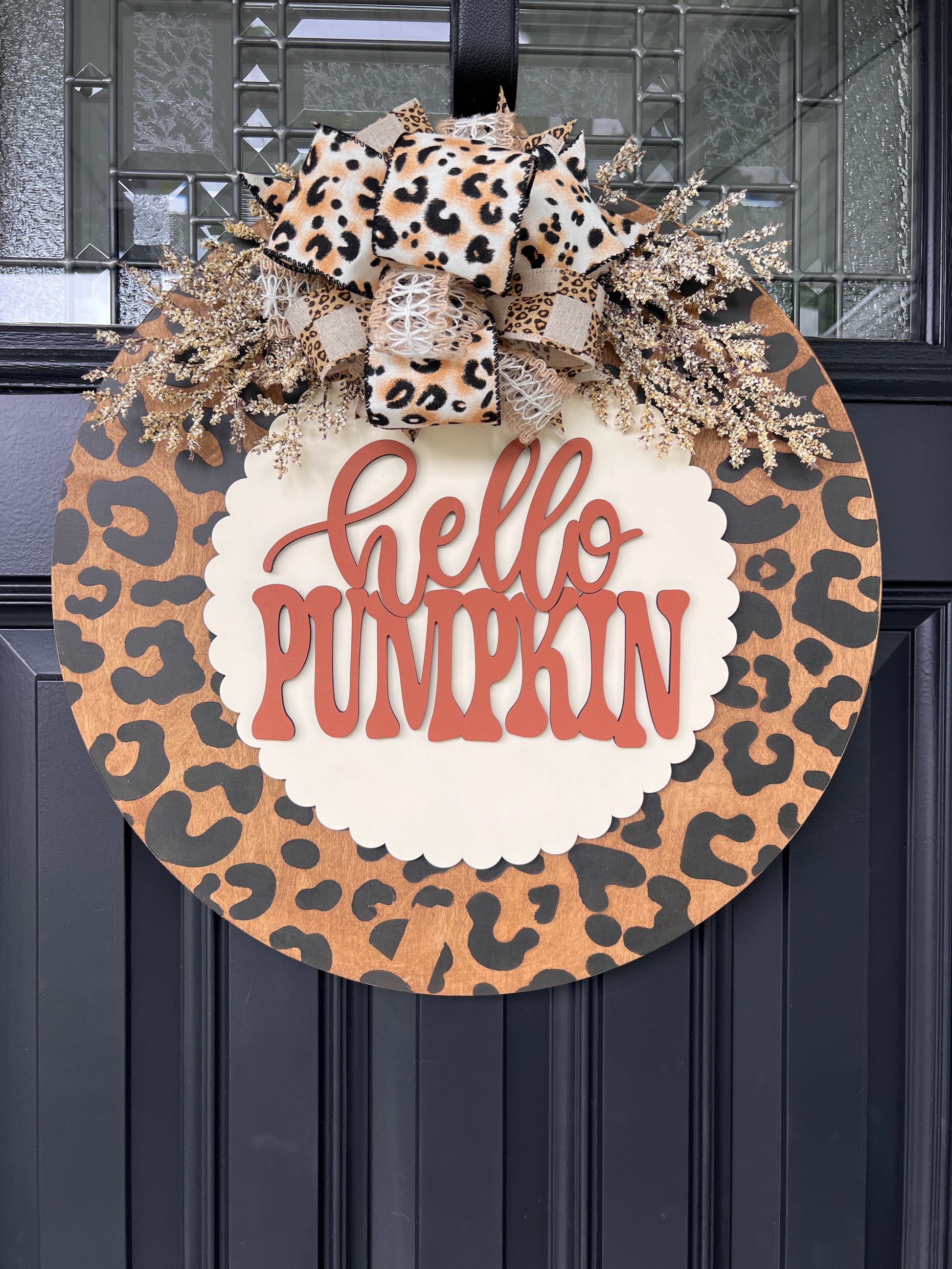 Hello Pumpkin leopard print fall door hanger with layered wood lettering, designer bow, seasonal greenery, and woodgrain animal print background displayed on a front door