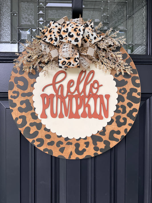 Hello Pumpkin leopard print fall door hanger with layered wood lettering, designer bow, seasonal greenery, and woodgrain animal print background displayed on a front door