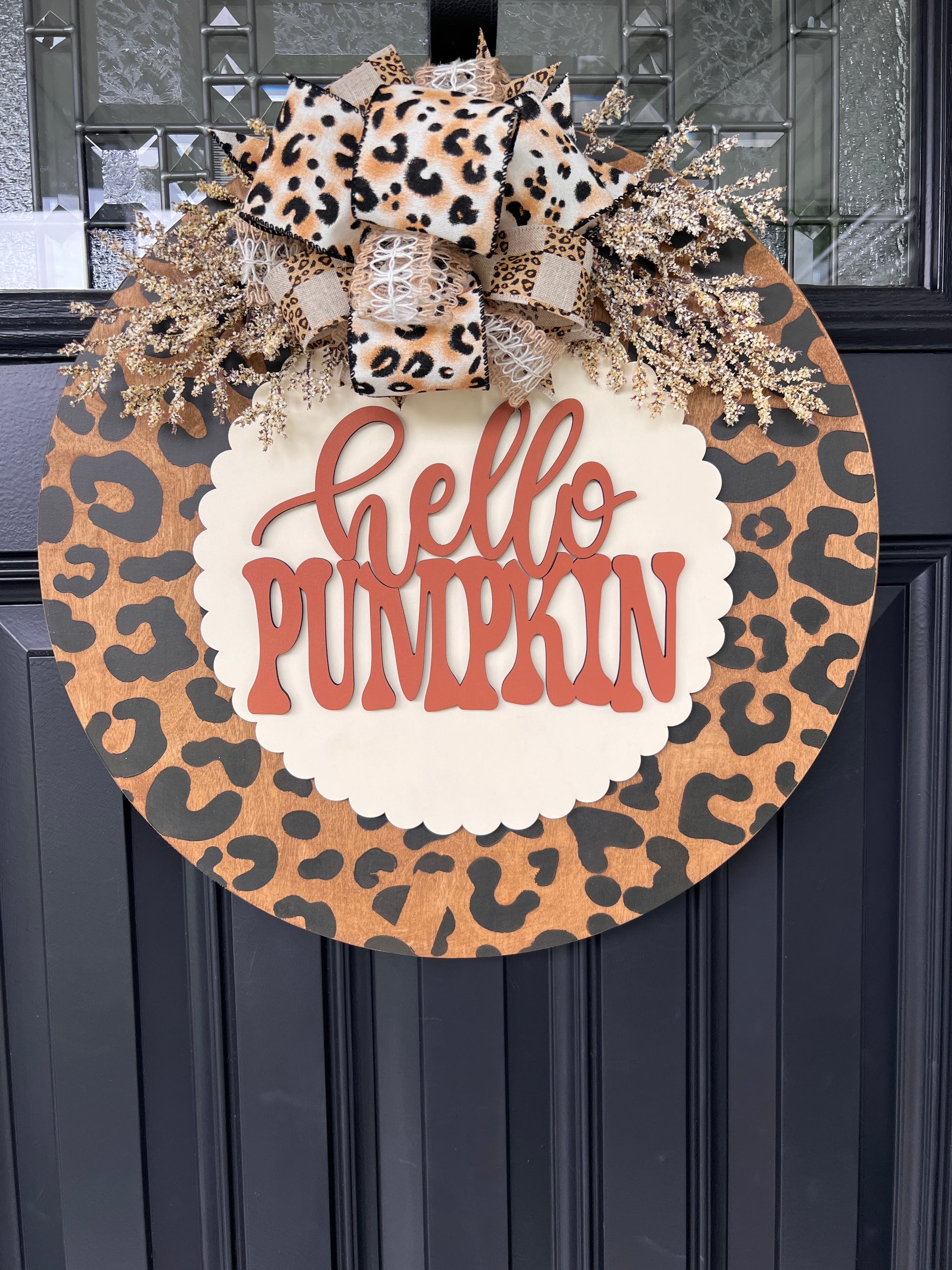 Hello Pumpkin leopard print fall door hanger with layered wood lettering, designer bow, seasonal greenery, and woodgrain animal print background displayed on a front door