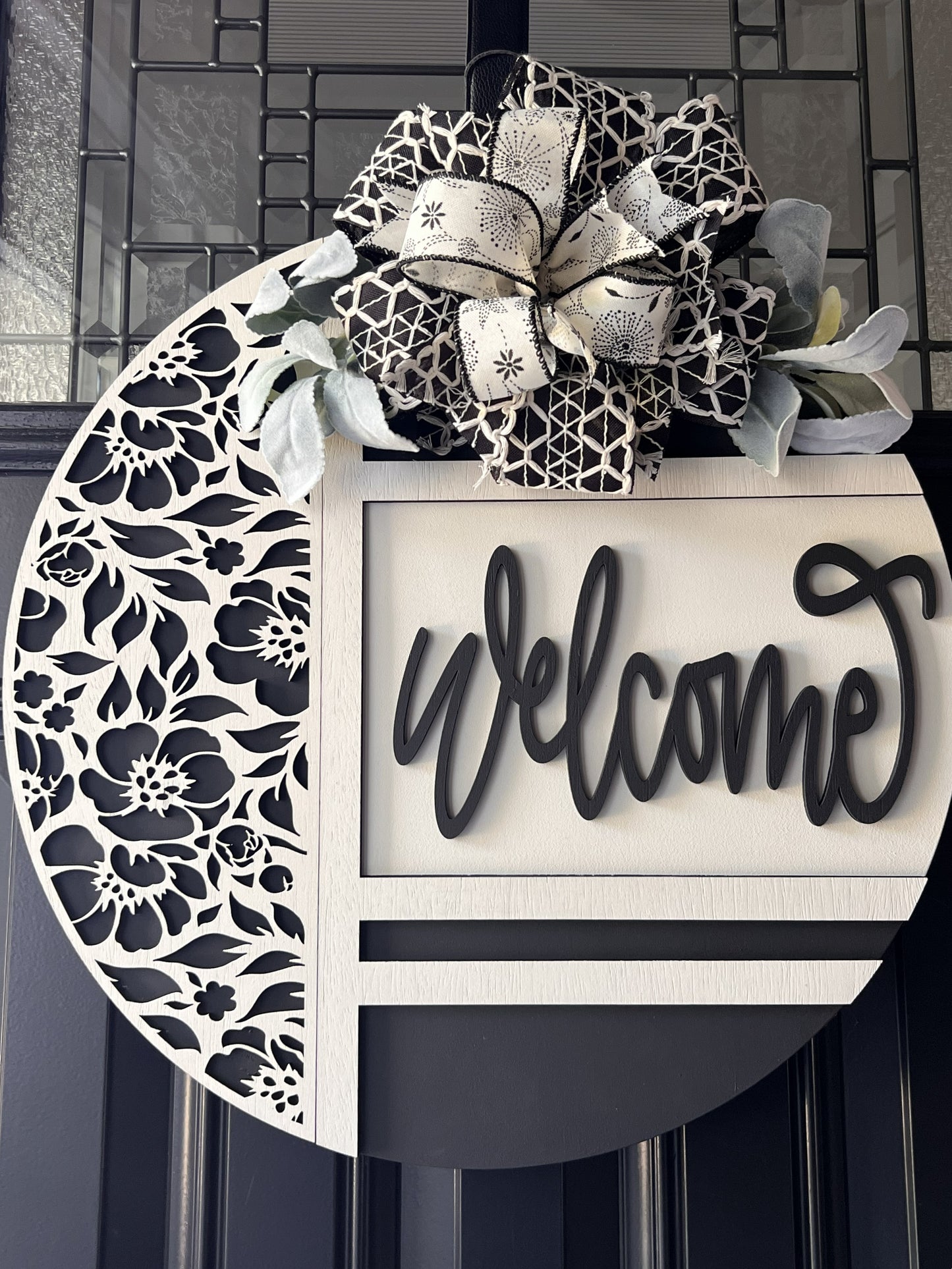 black and white welcome door hanger with floral cutout design
modern farmhouse front door sign with layered wood welcome script
18 inch round wooden welcome sign with decorative bow and greenery
neutral black white door hanger for front porch decor
handmade welcome sign with floral overlay and raised lettering
elegant entryway decor black and white round door hanger