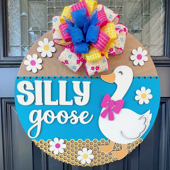 Handcrafted Silly Goose door hanger featuring a white goose with a pink bow, layered 3D lettering, colorful daisy accents, blue background, honeycomb detail, and decorative bow for farmhouse front door decor.
