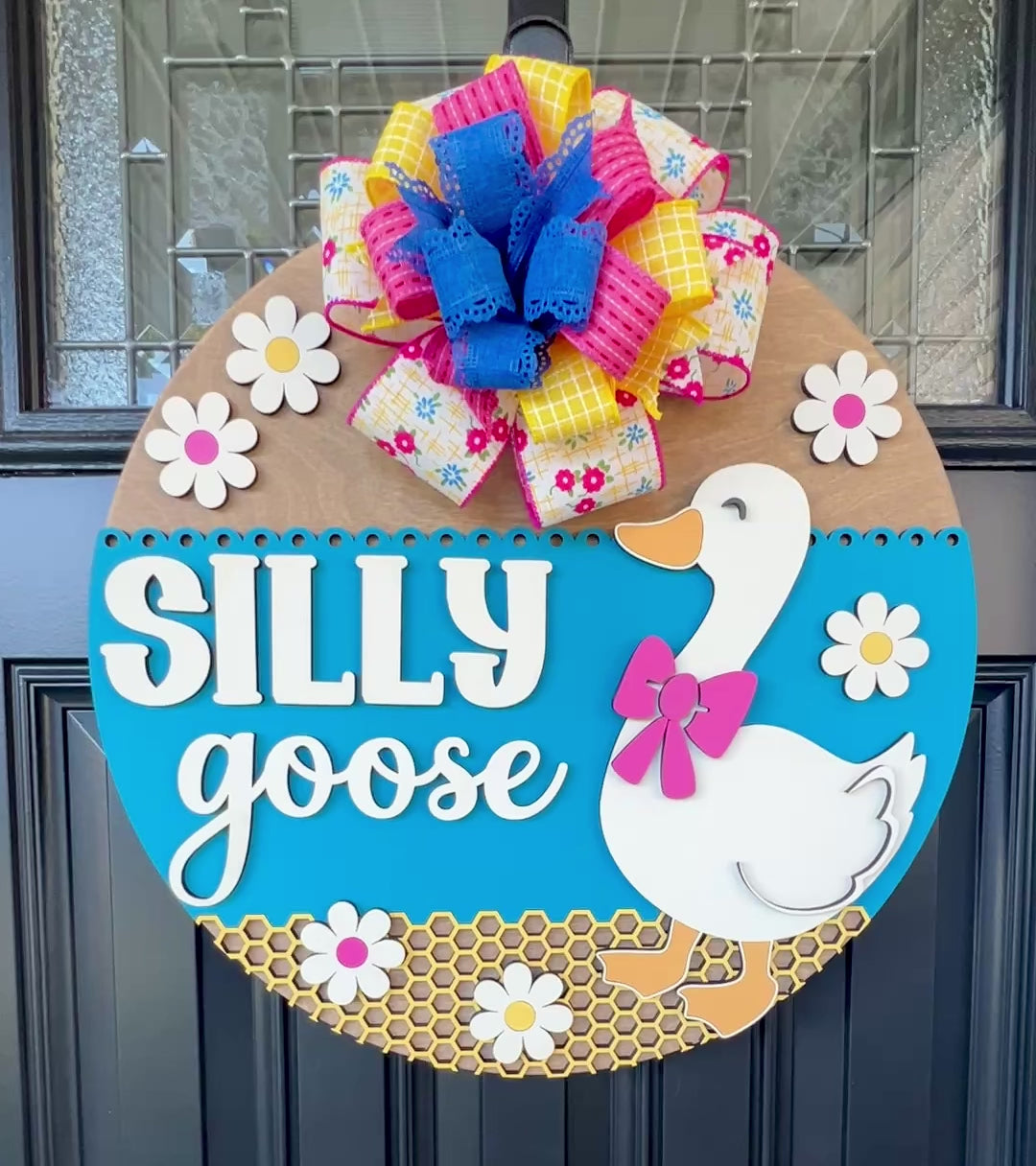 Handcrafted Silly Goose door hanger featuring a white goose with a pink bow, layered 3D lettering, colorful daisy accents, blue background, honeycomb detail, and decorative bow for farmhouse front door decor.