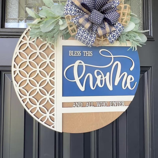 Navy blue and natural wood Bless This Home door hanger with layered 3D lettering, geometric overlay design, greenery accents, and designer bow displayed on a black front door.