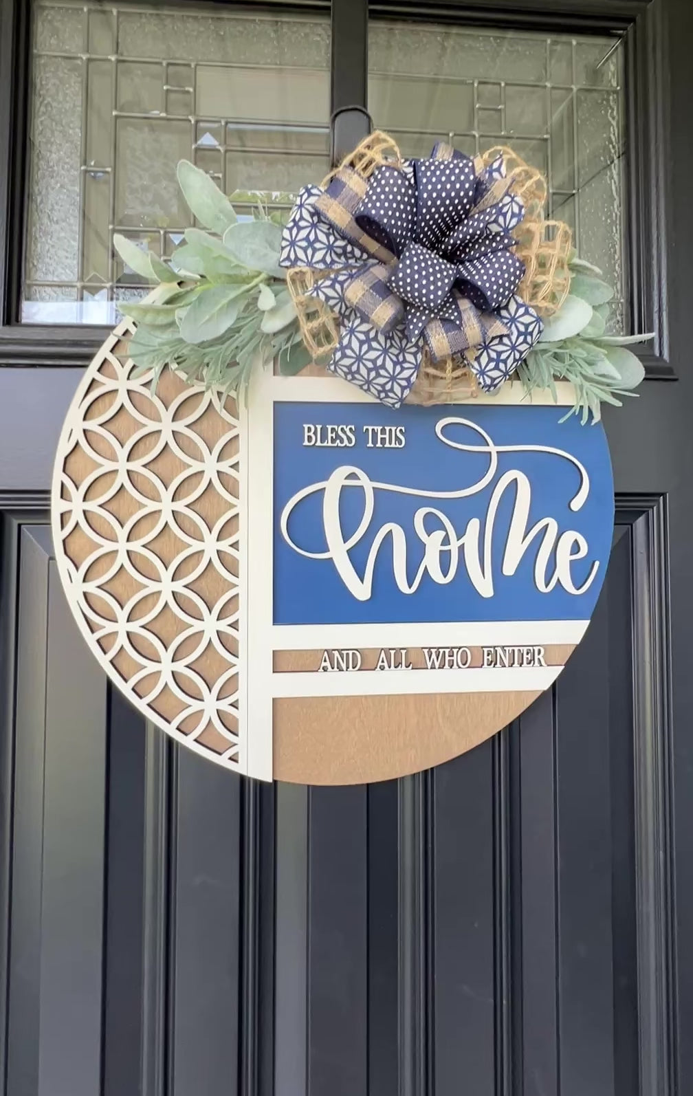 Navy blue and natural wood Bless This Home door hanger with layered 3D lettering, geometric overlay design, greenery accents, and designer bow displayed on a black front door.