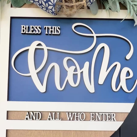 Navy blue and natural wood Bless This Home door hanger with layered 3D lettering, geometric overlay design, greenery accents, and designer bow displayed on a black front door.