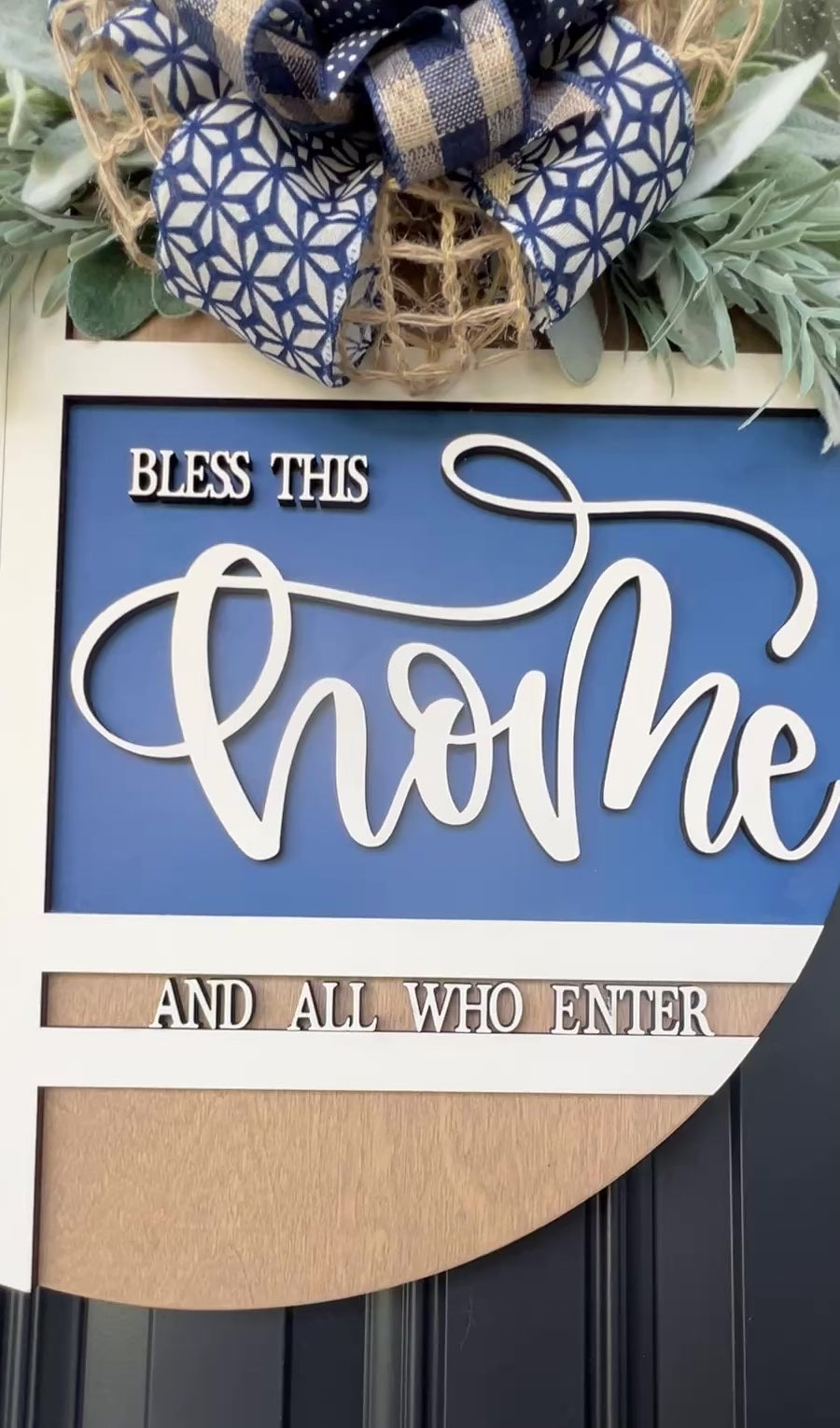 Navy blue and natural wood Bless This Home door hanger with layered 3D lettering, geometric overlay design, greenery accents, and designer bow displayed on a black front door.