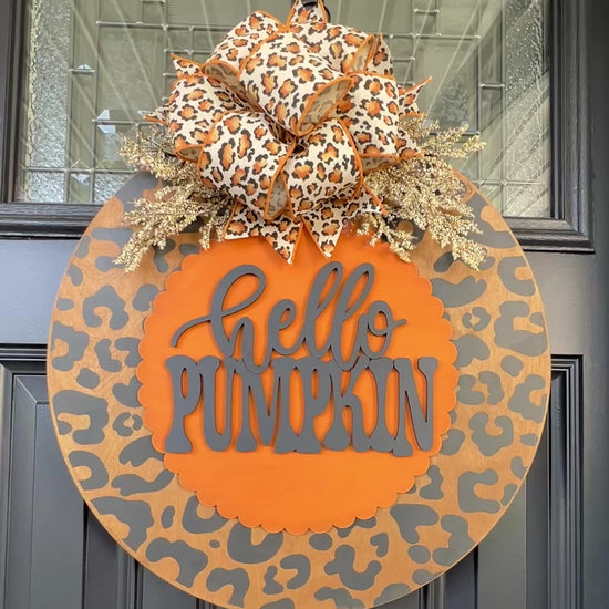 Hello Pumpkin leopard print fall door hanger with layered wood lettering, designer bow, seasonal greenery, and woodgrain animal print background displayed on a front door