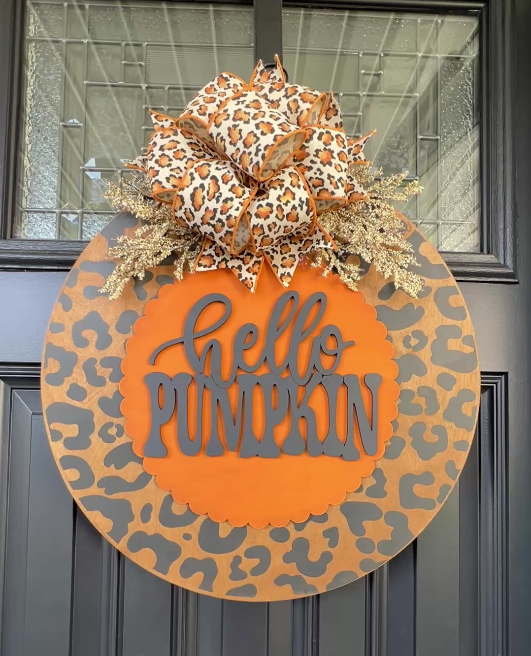 Hello Pumpkin leopard print fall door hanger with layered wood lettering, designer bow, seasonal greenery, and woodgrain animal print background displayed on a front door