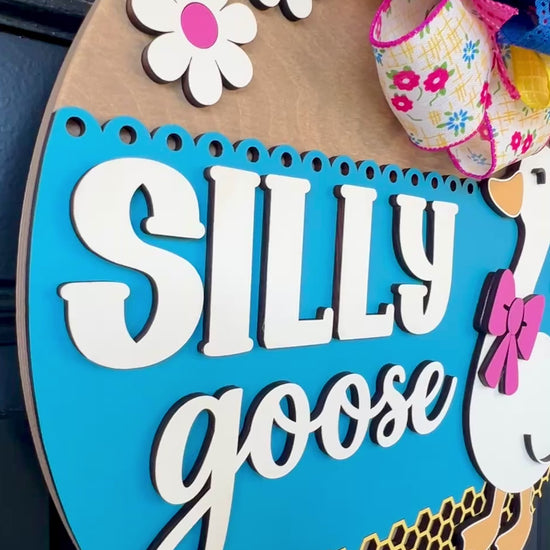 Handcrafted Silly Goose door hanger featuring a white goose with a pink bow, layered 3D lettering, colorful daisy accents, blue background, honeycomb detail, and decorative bow for farmhouse front door decor.
