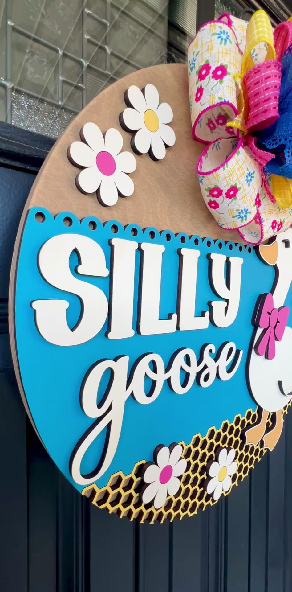 Handcrafted Silly Goose door hanger featuring a white goose with a pink bow, layered 3D lettering, colorful daisy accents, blue background, honeycomb detail, and decorative bow for farmhouse front door decor.