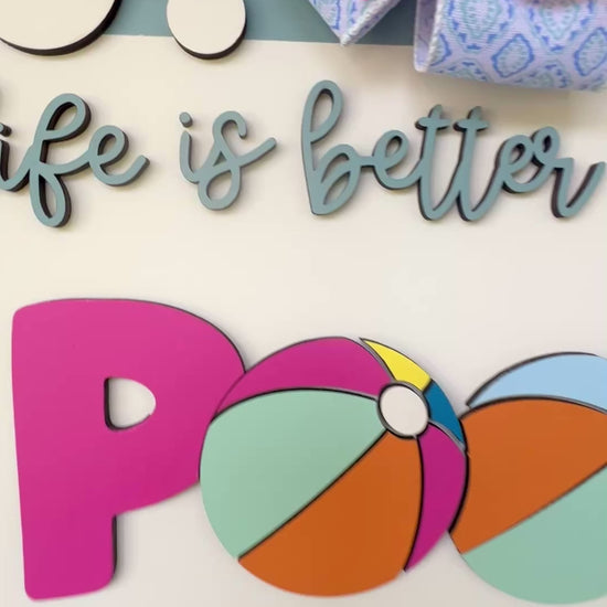 Handcrafted round door hanger featuring the phrase "Life Is Better at the Pool" with colorful beach ball accents, summer colors, and a bright pink and blue designer bow displayed on a front door.