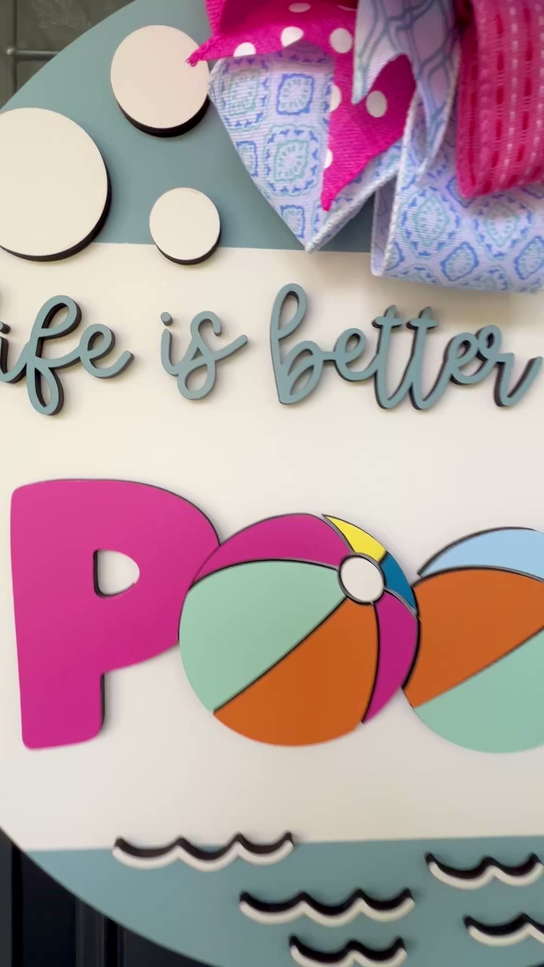 Handcrafted round door hanger featuring the phrase "Life Is Better at the Pool" with colorful beach ball accents, summer colors, and a bright pink and blue designer bow displayed on a front door.