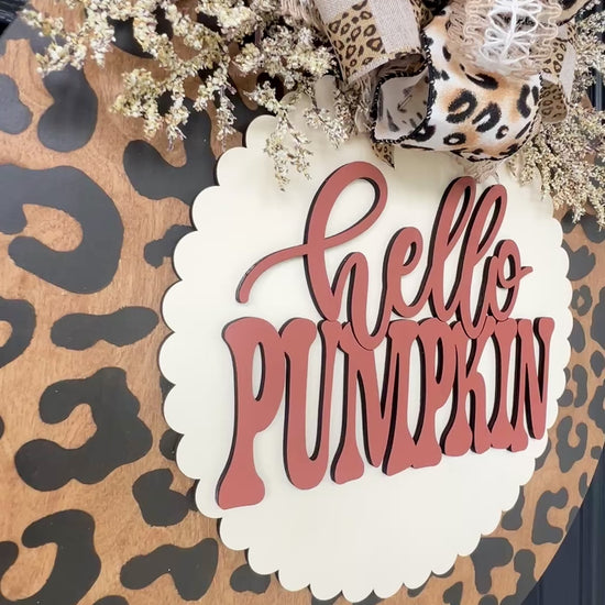 Hello Pumpkin leopard print door hanger with layered 3D lettering, designer bow, seasonal greenery, and woodgrain animal print background displayed on a front door