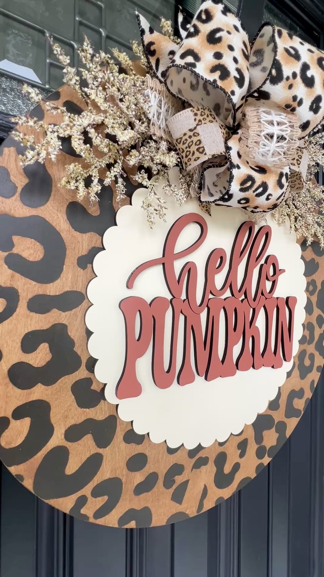 Hello Pumpkin leopard print door hanger with layered 3D lettering, designer bow, seasonal greenery, and woodgrain animal print background displayed on a front door