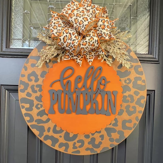 Hello Pumpkin leopard print fall door hanger with layered wood lettering, designer bow, seasonal greenery, and woodgrain animal print background displayed on a front door