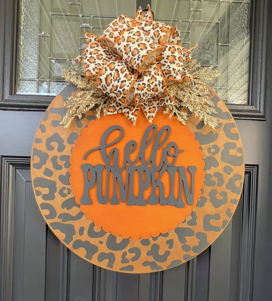Hello Pumpkin leopard print fall door hanger with layered wood lettering, designer bow, seasonal greenery, and woodgrain animal print background displayed on a front door